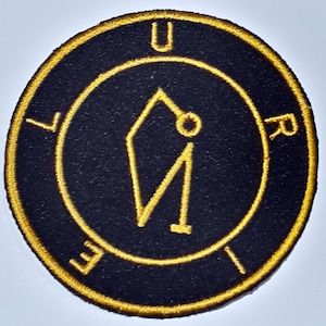 Peut inclure: Un écusson brodé noir et or avec un symbole stylisé et les lettres "URIE" autour.