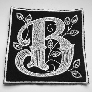 Op de afbeelding: Witte geborduurde letter B op een zwarte stoffen patch. De letter is in een decoratief lettertype met bloemmotieven.