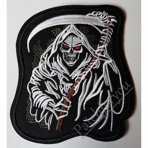 以下が含まれることがあります： 黒い刺繍パッチに、白い骨格の図形が鎌を持っています。図形は赤い目をしており、フード付きのローブを着ています。背景は濃い緑色です。