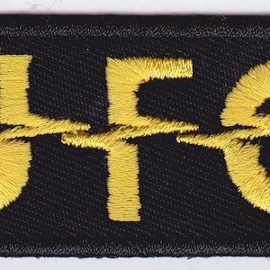 Könnte beinhalten: Schwarzer gestickter Aufnäher mit dem Wort "UFO" in gelbem Garn. Das "U" und das "O" sind mit einem Blitz durch die Mitte stilisiert.