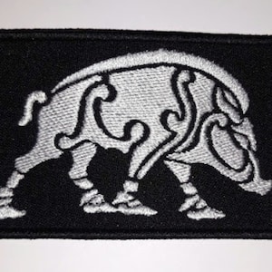 Puede incluir: Un parche bordado negro con un diseño de jabalí estilizado blanco. El jabalí está mirando hacia la derecha y tiene un diseño detallado con líneas y curvas intrincadas.