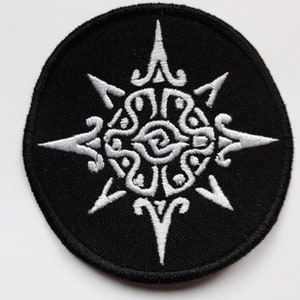 Peut inclure: Un écusson brodé blanc sur fond noir. Le motif est un soleil stylisé à huit pointes, chaque pointe se terminant par une pointe acérée. Le centre du soleil est rempli d'un motif tourbillonnant.