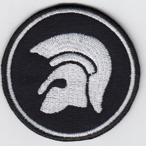 Puede incluir: Un parche redondo negro con un diseño de casco troyano blanco bordado. El casco está centrado dentro del parche, y el diseño está delineado con un borde blanco. Ideal para ropa o accesorios.