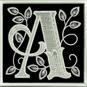 Op de afbeelding: Een witte geborduurde letter A met een bloemmotief op een zwarte achtergrond.