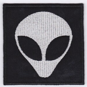 Op de afbeelding: Zwart vierkant geborduurd embleem met een wit alienhoofd. De alien heeft grote, ovale ogen en een gladde kop. Dit embleem is bedoeld om op kleding of accessoires te naaien of te strijken.