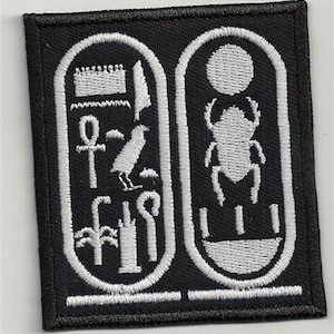 以下が含まれることがあります： 黒い布のパッチに白い刺繍で描かれたエジプトのヒエログリフ。ヒエログリフには、スカラベ、アンク、鳥、植物が含まれています。