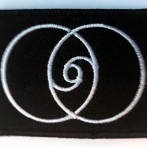 Op de afbeelding: Een zwarte geborduurde patch met een wit ontwerp van twee overlappende cirkels, waarvan er één een spiraal is.
