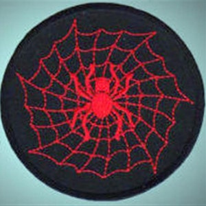 Op de afbeelding: Een zwarte stoffen patch met een rood geborduurd spinnenweb en spin in het midden.