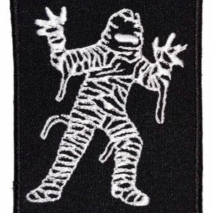 Peut inclure: Un écusson rectangulaire noir avec un motif de momie blanche. La momie est représentée avec des bandelettes et des bras tendus, donnant un aspect effrayant. L'écusson est destiné aux vêtements ou accessoires.