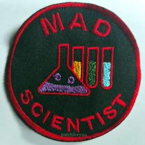 Puede incluir: Un parche redondo con borde rojo sobre fondo negro. El parche presenta el texto "MAD SCIENTIST" en bordado rojo. Un vaso de precipitados morado, rojo y amarillo se encuentra en el centro del parche, con tres tubos de ensayo llenos de líquido amarillo, verde y azul.