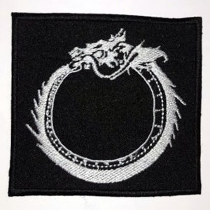Pode incluir: Etiqueta bordada preta com um dragão branco enroscado em um círculo. A cabeça do dragão está voltada para o observador e sua cauda está enroscada ao redor do círculo. O círculo tem uma borda branca com um design.