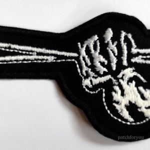May include: Black embroidered patch featuring a white skeleton hand holding two drumsticks. The patch is shaped like a hand pointing to the right.