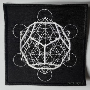 Puede incluir: Parche de tela negro con un diseño geométrico bordado en blanco. El diseño presenta un cubo con círculos superpuestos a su alrededor.
