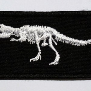 Może przedstawiać: Prostokątna czarna naszywka z białym haftowanym wzorem szkieletu Tyrannosaurus Rex. Szczegółowe szwy podkreślają strukturę kości dinozaura. Idealna do personalizacji odzieży i akcesoriów.