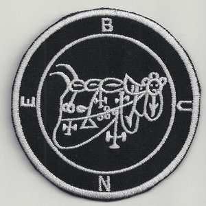 May include: A black and white embroidered patch with a circular design. The center of the patch features a stylized image of a demon with a compass rose around it. The compass rose is labeled with the letters N, S, E, and W. The demon is surrounded by a circle with the letters B, C, and N.