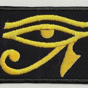 Könnte beinhalten: Ein schwarzer gestickter Aufnäher mit einem gelben Horusauge-Symbol.