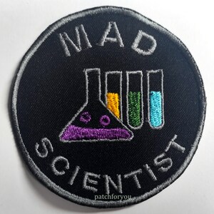 Puede incluir: Un parche bordado negro y gris con el texto "MAD SCIENTIST" en un diseño circular. El parche presenta un vaso de precipitados y tubos de ensayo morados, amarillos, verdes y azules.