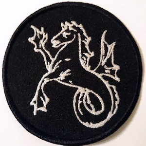 Pode incluir: Um remendo circular preto com uma imagem bordada branca de uma criatura mítica de cavalo-marinho com asas.