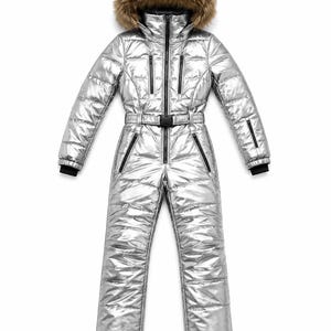 Op de afbeelding: Een metallic zilveren skipak uit één stuk met een gewatteerd ontwerp, een ritssluiting aan de voorzijde en een tailleband met een zwarte gesp. De capuchon is afgezet met bruin imitatiebont en de mouwen hebben zwarte boorden.