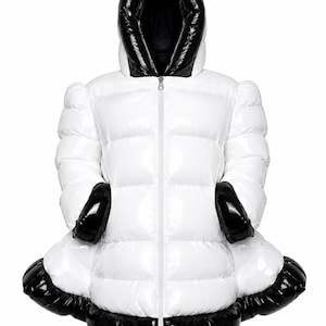 Può includere: Un piumino bianco con finitura lucida, caratterizzato da una fodera del cappuccio nera, polsini neri e un orlo nero. La giacca presenta una chiusura centrale con zip, pannelli trapuntati orizzontali e una silhouette svasata.