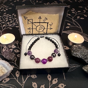 May include: A beaded bracelet featuring black, purple, and dark pink spherical beads on a black cord, presented in a black box with a white interior. The box lid displays a tan paper insert with a black ink drawing of a geometric symbol.