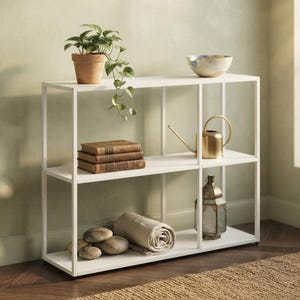 May include: A white metal bookshelf with three horizontal tiers and a rectangular frame. The shelves hold a potted green plant, a ceramic bowl, three stacked brown books, a gold watering can, a metal lantern, and a rolled beige fabric cloth.