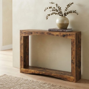 May include: A rectangular console table with a dark brown wood grain finish, featuring a curved top and bottom shelf design. A two-toned ceramic vase with dried eucalyptus branches and a book titled Artisanal Homes sit on the table surface.