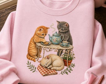 Op de afbeelding: Een lichtroze sweater met ronde hals en een afbeelding van drie kittens. Een oranje kitten zit aan een tafel met een theepot en theekopje, terwijl een grijs kitten ernaast staat. Een crèmekleurig kitten slaapt op een geruit kleedje.