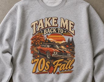 Op de afbeelding: Een grijs gemêleerd sweatshirt met ronde hals en een grafische print van een vintage oranje sportwagen op een weg omringd door herfstbomen. De tekst "TAKE ME BACK TO 70s Fall" staat in een retro lettertype met een zonsondergang op de achtergrond.
