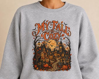Op de afbeelding: Een grijs gemêleerd sweatshirt met ronde hals en een grafische print met de tekst "MY FALL IS COZY" in het oranje. De illustratie toont een griezelig dorpsgezicht bij nacht met een volle maan, kale bomen, een zwarte kat en pompoenen.