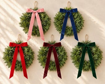 May include: Five circular evergreen wreaths arranged in a group, each featuring a long velvet ribbon tied in a bow at the top. The ribbons are coloured in shades of light pink, navy blue, bright red, deep burgundy, and dark forest green.