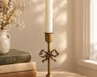 May include: A gold-toned metal candlestick holder featuring a decorative bow detail on the central stem and a fluted circular base. A single white taper candle is inserted into the top cup of the holder, which stands upright on a flat surface.