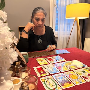 Puede incluir: Un conjunto de cartas de tarot dispuestas en un despliegue sobre un mantel rojo. Las cartas presentan varias ilustraciones coloridas y texto. Varios portavelas de cristal peque&ntilde;os con velas de t&eacute; est&aacute;n colocados cerca de las cartas.