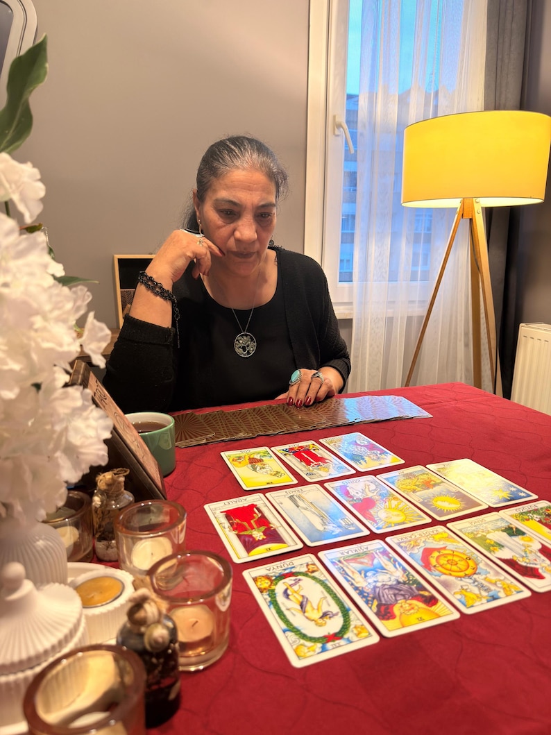 Puede incluir: Un conjunto de cartas de tarot dispuestas en un despliegue sobre un mantel rojo. Las cartas presentan varias ilustraciones coloridas y texto. Varios portavelas de cristal peque&ntilde;os con velas de t&eacute; est&aacute;n colocados cerca de las cartas.
