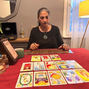 Puede incluir: Un conjunto de cartas del tarot dispuestas en un despliegue sobre un mantel rojo. Las cartas presentan ilustraciones y s&iacute;mbolos coloridos. Una caja de madera etiquetada como Tarot se encuentra a la izquierda, junto a portavelas de cristal y una taza verde.