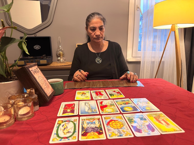 Puede incluir: Un conjunto de cartas del tarot dispuestas en un despliegue sobre un mantel rojo. Las cartas presentan ilustraciones y s&iacute;mbolos coloridos. Una caja de madera etiquetada como Tarot se encuentra a la izquierda, junto a portavelas de cristal y una taza verde.