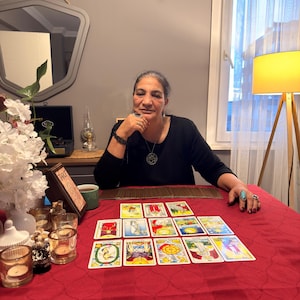 Puede incluir: Un conjunto de cartas del tarot dispuestas en una cuadr&iacute;cula sobre un mantel rojo. Las cartas presentan ilustraciones coloridas y texto. Un peque&ntilde;o letrero de madera con la palabra Tarot se coloca cerca de una taza verde, flores y velas.