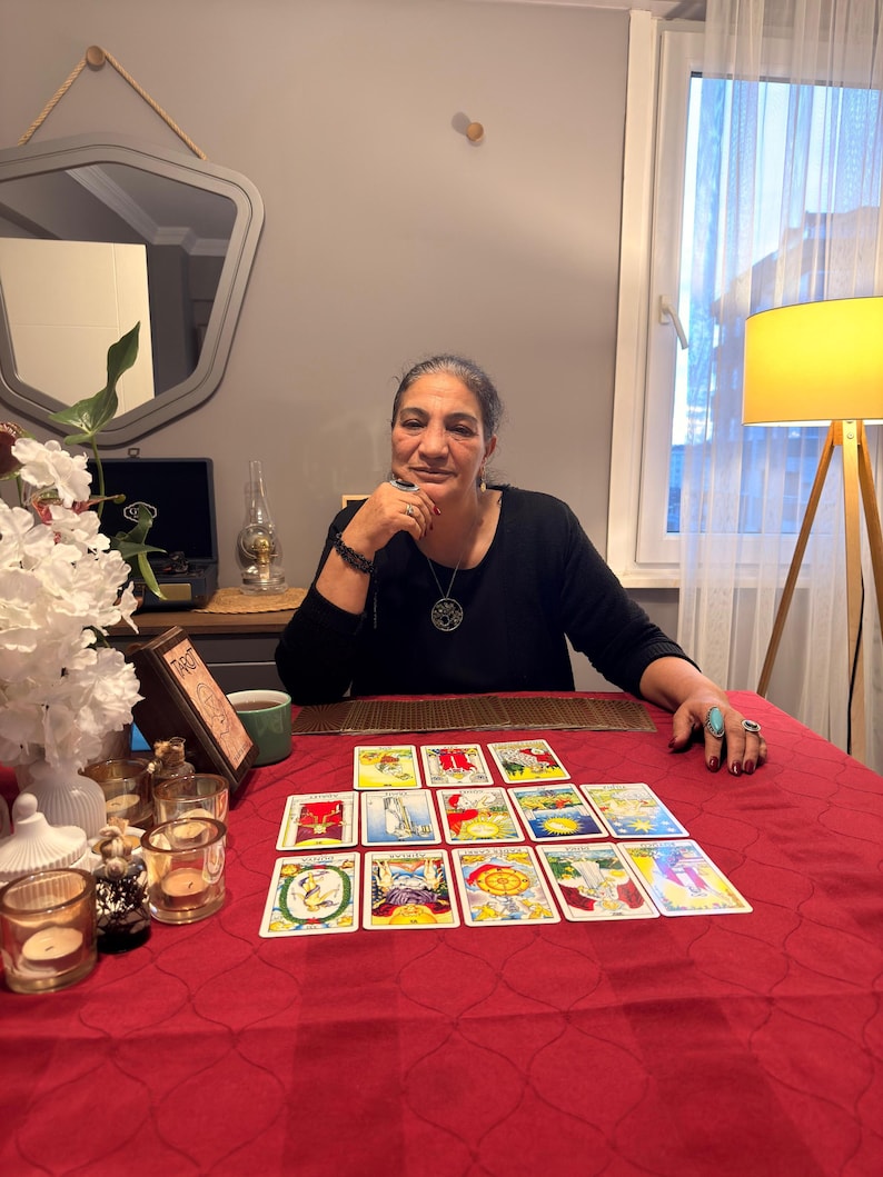 Puede incluir: Un conjunto de cartas del tarot dispuestas en una cuadr&iacute;cula sobre un mantel rojo. Las cartas presentan ilustraciones coloridas y texto. Un peque&ntilde;o letrero de madera con la palabra Tarot se coloca cerca de una taza verde, flores y velas.