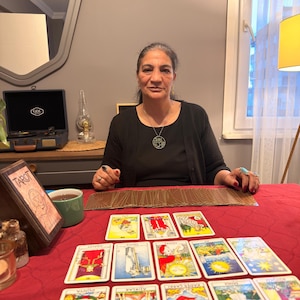 Puede incluir: Una persona con una blusa negra y un collar con colgante de &aacute;rbol circular se sienta tras una mesa roja con cartas de tarot extendidas. Se ven una caja de madera con la palabra Tarot, una taza verde y un tocadiscos antiguo sobre la mesa.