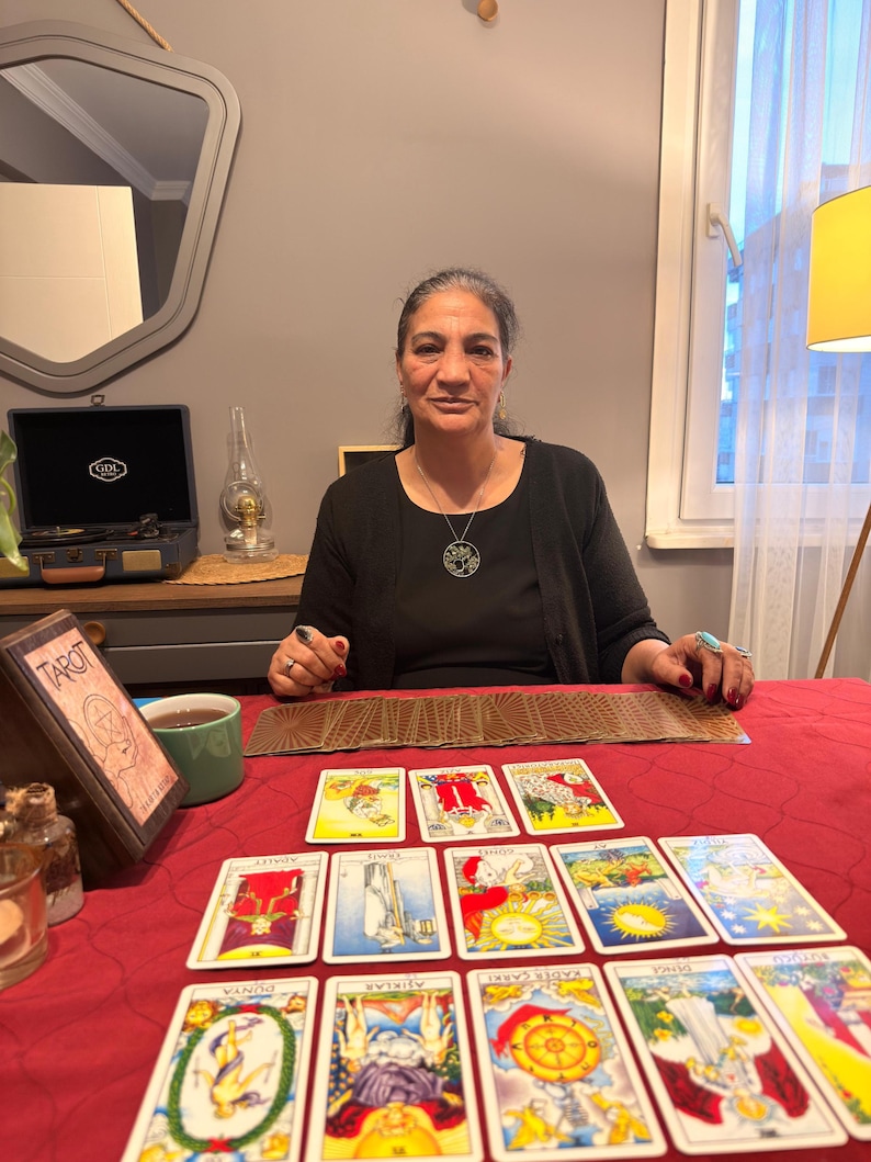 Puede incluir: Una persona con una blusa negra y un collar con colgante de &aacute;rbol circular se sienta tras una mesa roja con cartas de tarot extendidas. Se ven una caja de madera con la palabra Tarot, una taza verde y un tocadiscos antiguo sobre la mesa.