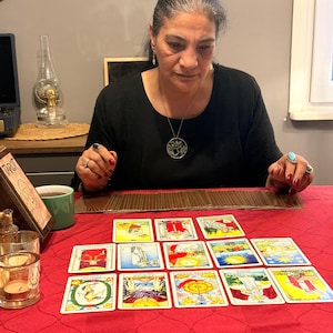 Puede incluir: Un conjunto de cartas de tarot dispuestas en un despliegue sobre un mantel rojo. Las cartas presentan ilustraciones coloridas y texto en turco. Un soporte de madera para cartas de tarot se encuentra a la izquierda, junto a dos portavelas de cristal y una taza verde.