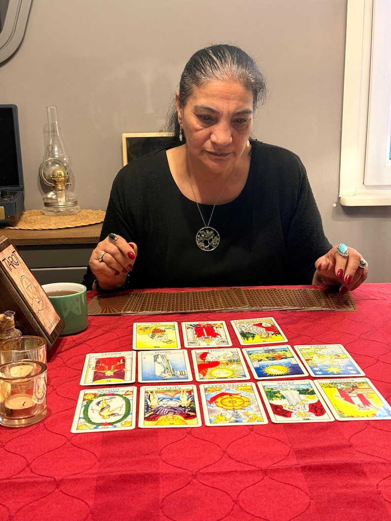 Puede incluir: Un conjunto de cartas de tarot dispuestas en un despliegue sobre un mantel rojo. Las cartas presentan ilustraciones coloridas y texto en turco. Un soporte de madera para cartas de tarot se encuentra a la izquierda, junto a dos portavelas de cristal y una taza verde.