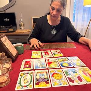 Puede incluir: Un conjunto de cartas de tarot dispuestas sobre un mantel rojo. Las cartas presentan varias ilustraciones coloridas y texto. Una caja de madera etiquetada como TAROT se encuentra a la izquierda, junto a una taza verde y dos portavelas de cristal.