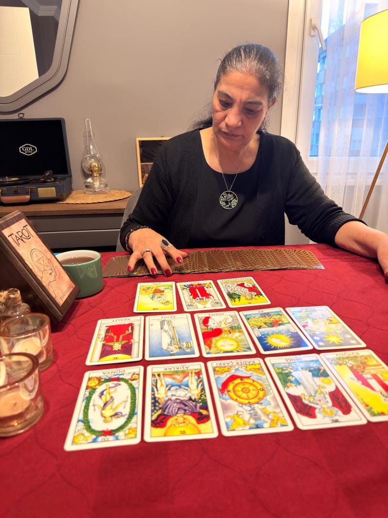 Puede incluir: Un conjunto de cartas de tarot dispuestas sobre un mantel rojo. Las cartas presentan varias ilustraciones coloridas y texto. Una caja de madera etiquetada como TAROT se encuentra a la izquierda, junto a una taza verde y dos portavelas de cristal.
