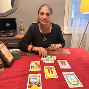 Puede incluir: Una persona con una camisa negra de manga larga y un collar con colgante circular est&aacute; sentada a una mesa con un mantel rojo. Varias cartas del tarot est&aacute;n dispuestas sobre la superficie, incluyendo AŞIKLAR, B&Uuml;Y&Uuml;C&Uuml; y D&Uuml;NYA, junto a una baraja.