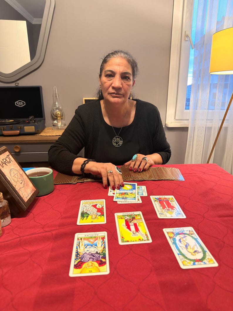Puede incluir: Una persona con una camisa negra de manga larga y un collar con colgante circular est&aacute; sentada a una mesa con un mantel rojo. Varias cartas del tarot est&aacute;n dispuestas sobre la superficie, incluyendo AŞIKLAR, B&Uuml;Y&Uuml;C&Uuml; y D&Uuml;NYA, junto a una baraja.