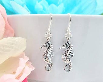 May include: A pair of silver dangle earrings featuring a detailed seahorse design. The earrings are hanging from a white surface.