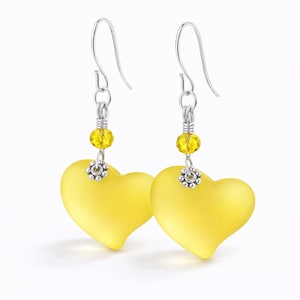 May include: A pair of heart-shaped earrings. Each earring features a matte yellow heart pendant suspended from a silver-colored hook. A small cluster of yellow beads and a decorative silver element are positioned above the heart.