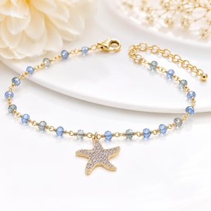 Puede incluir: Una pulsera de cadena dorada con cuentas facetadas en azul claro y transparente. La pulsera incluye un dije central de estrella de mar dorada incrustado con pequeñas piedras transparentes y un cierre de mosquetón con una cadena de extensión.