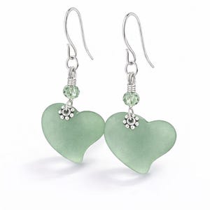 May include: A pair of heart-shaped earrings. Each earring features a pale green, frosted glass heart pendant. The pendants are connected to silver-colored ear wires with a small green bead and a decorative silver-colored flower.